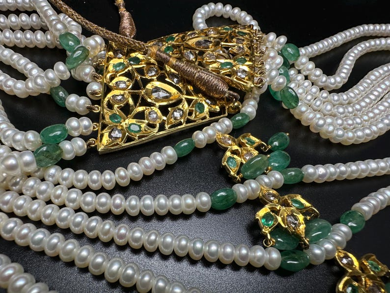 Puede incluir: Un collar ornamentado con m&uacute;ltiples hilos de perlas blancas, intercaladas con piedras preciosas verdes. Un colgante dorado con detalles intrincados y gemas incrustadas es el punto focal. El collar se presenta sobre un fondo oscuro.