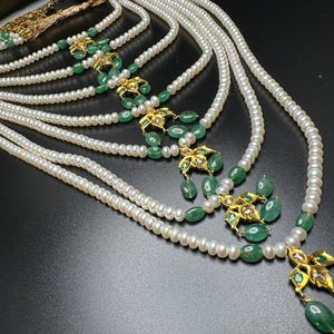 Puede incluir: Un collar de perlas de m&uacute;ltiples hilos con detalles de esmeraldas y oro. El collar presenta m&uacute;ltiples hilos de peque&ntilde;as perlas blancas, intercaladas con dise&ntilde;os florales dorados y cuentas de esmeralda verde. El collar se exhibe sobre una superficie oscura.