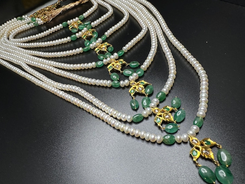 Puede incluir: Un collar de perlas de m&uacute;ltiples hilos con detalles de esmeraldas y oro. El collar presenta m&uacute;ltiples hilos de peque&ntilde;as perlas blancas, intercaladas con dise&ntilde;os florales dorados y cuentas de esmeralda verde. El collar se exhibe sobre una superficie oscura.
