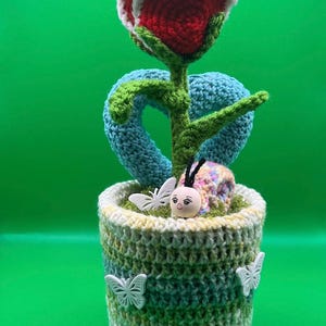 Può includere: Una composizione floreale all'uncinetto fatta a mano con una rosa rossa e bianca, uno stelo verde e foglie blu a forma di cuore. Il fiore è in un vaso con accenti di farfalla bianca e un bruco decorativo.