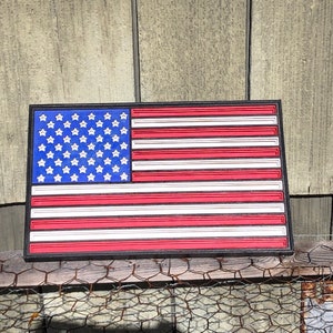 May include: A 3D printed American flag with red, white, and blue stars and stripes. The flag is framed in black and is hanging on a wooden fence.