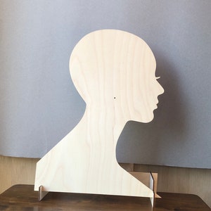 May include: A wooden silhouette of a person's head and shoulders, standing on a small wooden base. The silhouette is made of light-colored wood and is unpainted.
