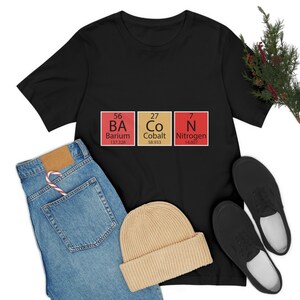 Könnte beinhalten: Ein schwarzes Kurzarm-T-Shirt mit einer Grafik aus drei Periodensystem-Elementen, die BACON buchstabieren: Barium (BA), Cobalt (Co) und Stickstoff (N). Das Design enthält Ordnungszahlen und Atommassen für jedes Element in roten und beigen Kästchen.