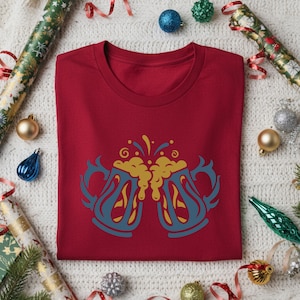 Puede incluir: Una camiseta roja con un diseño estilizado de dos jarras de cerveza con asas azules y espuma dorada. Las jarras chocan entre sí, con un tema festivo. La camiseta está doblada sobre una superficie blanca con adornos navideños.