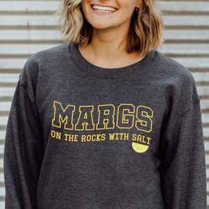 May include: A dark gray sweatshirt with the text "MARGS ON THE ROCKS WITH SALT" in yellow lettering. A yellow lemon slice graphic is below the text.