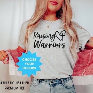 May include: Athletic heather grey premium tee with the text "Raising Warriors" in black script, featuring a heart and cross design. A blue starburst graphic reads "Choose Your Colors".
