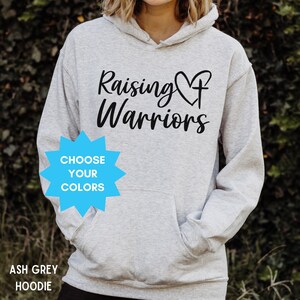 May include: A light grey hoodie featuring the words "Raising Warriors" in black script. The hoodie has a front pocket and a drawstring hood. A blue starburst graphic reads "Choose Your Colors".