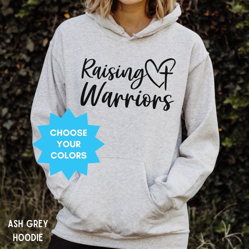 May include: A light grey hoodie featuring the words "Raising Warriors" in black script. The hoodie has a front pocket and a drawstring hood. A blue starburst graphic reads "Choose Your Colors".