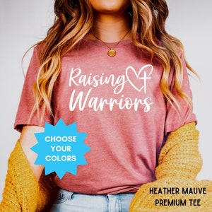 May include: Heather mauve premium tee with the words "Raising Warriors" in white script. A heart and cross design is incorporated into the text. A blue starburst graphic reads "Choose Your Colours".