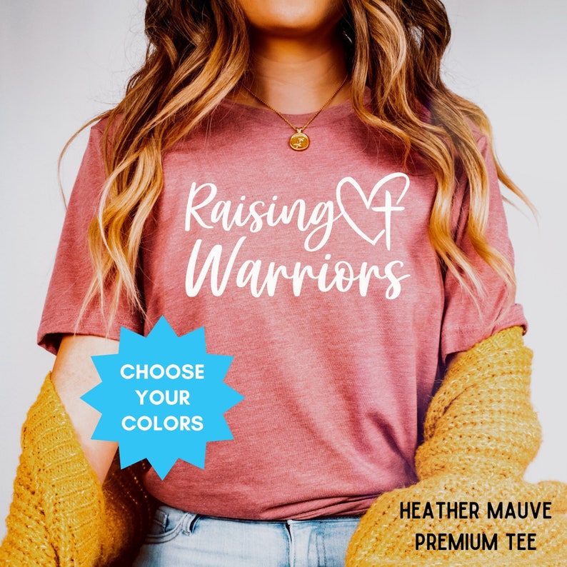 May include: Heather mauve premium tee with the words "Raising Warriors" in white script. A heart and cross design is incorporated into the text. A blue starburst graphic reads "Choose Your Colours".