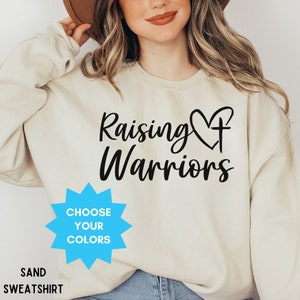 May include: A sand-coloured sweatshirt with the words "Raising Warriors" in black script, featuring a heart and cross design. A blue starburst graphic reads "Choose Your Colors." The bottom of the sweatshirt says "Sand Sweatshirt."