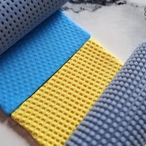 Può includere: Due lastre di pasta polimerica testurizzata blu e gialla con un rullo testurizzato grigio su ogni lato. Il rullo sul lato destro è parzialmente sulla lastra gialla. Il testo "Grumpy Guppy clay tools" è nell'angolo in alto a destra dell'immagine.