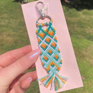 May include: A green, white, and orange friendship bracelet with a heart pattern. The bracelet is attached to a silver key chain.