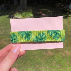 May include: A green and yellow friendship bracelet with a repeating pattern of green leaves. The bracelet is displayed on a pink card.