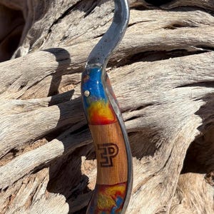 Peut inclure: Un outil crochet en métal courbé avec un manche composé d'une section centrale en bois clair portant un logo marron foncé avec les lettres P et P, entourée de sections en résine tourbillonnante bleue, rouge et dorée, fixées par deux goupilles.