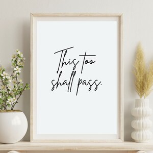 May include: A framed print with the text "This too shall pass" in black cursive script on a white background. The print is displayed on a light wood shelf with a white vase and a bouquet of white flowers on the left and a vase with dried beige grass on the right.