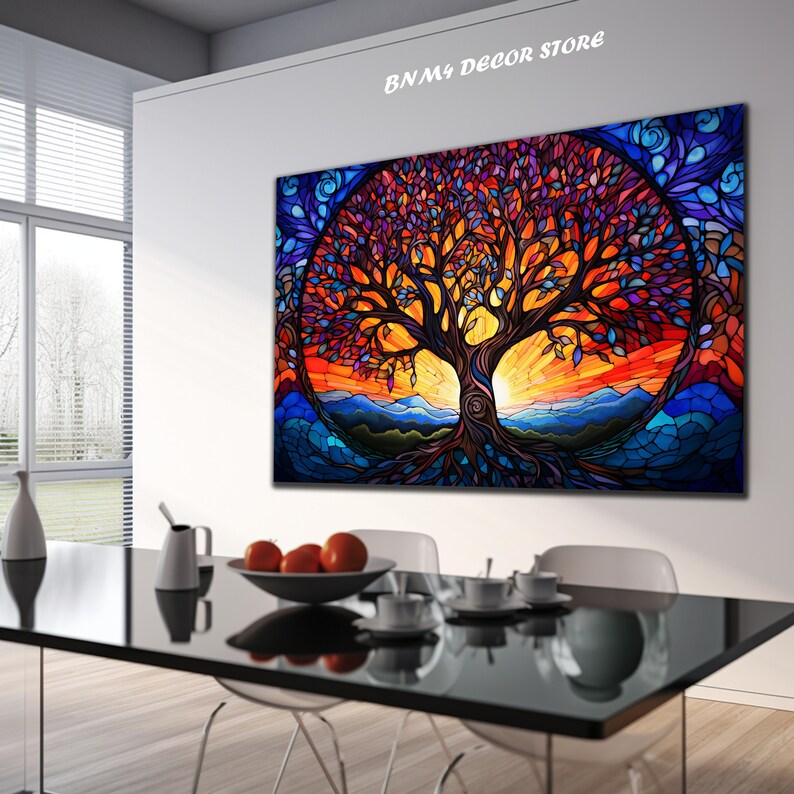 May include: A colorful stained glass style painting of a tree with a sunset in the background. The tree has a thick trunk and many branches with leaves in shades of purple, blue, orange, and yellow. The sunset is a vibrant orange and red with mountains in the distance. The painting is framed and hanging on a white wall in a room with a dining table and chairs.