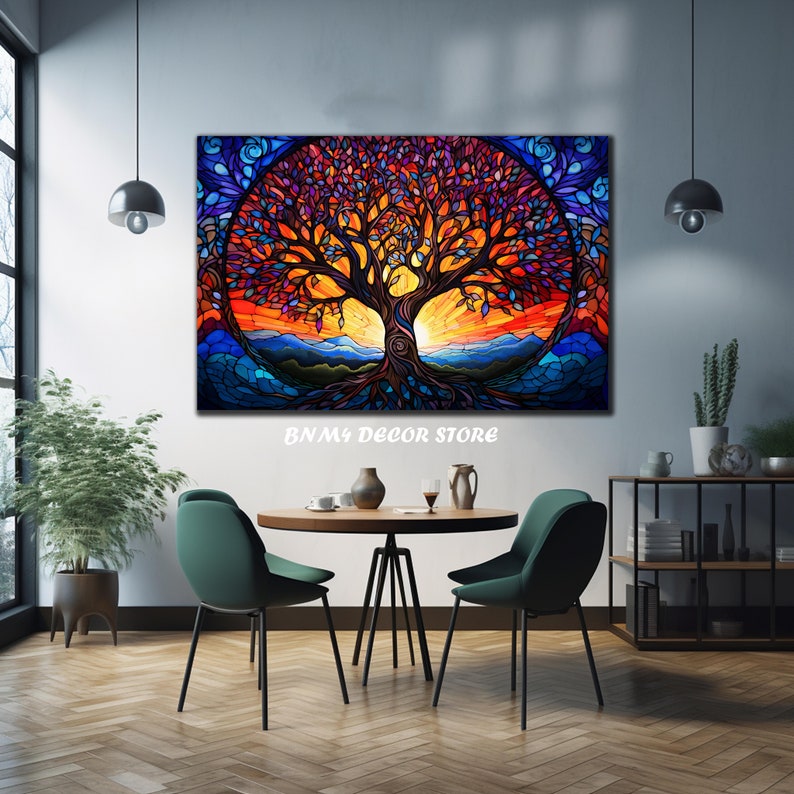 May include: A colorful stained glass style painting of a tree with a sunset in the background. The tree has a thick trunk and many branches with leaves in shades of purple, orange, and blue. The sunset is a vibrant orange and yellow with a blue sky above. The painting is framed and hanging on a white wall in a room with a table and chairs.