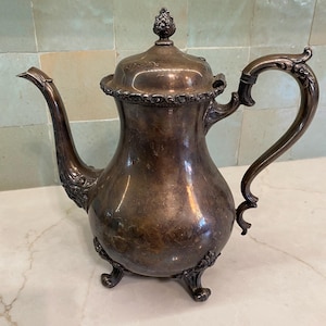 May include: A tarnished silver-toned metal teapot featuring a curved spout, a decorative handle, and a lid topped with a finial. The body of the teapot rests on three ornate feet and displays intricate embossed floral detailing along the edges.