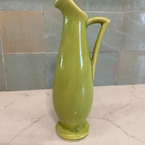 May include: A tall, lime green ceramic pitcher with a smooth, glossy finish. The vessel features a slender, elongated body, a flared pouring spout, a curved handle attached to the side, and a circular base that provides stability for the upright form.