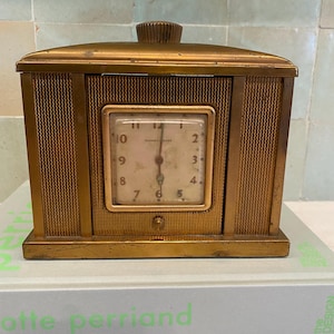 May include: A vintage gold-toned travel clock with a rectangular face and a square clock face. The clock has a mesh-like design on the sides and a small knob on top. The clock face displays the time, with the hands pointing to approximately 6:00.