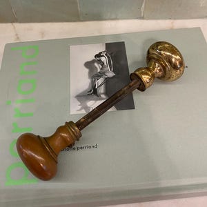 May include: A vintage door handle with a brass finish on the oval-shaped knobs. The handle has a wooden grip and a threaded rod. The handle is resting on a book with a black and white image of a woman.