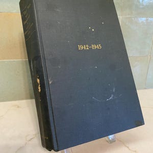 May include: A vintage hardcover book with a dark blue cover. The title "1942-1945" is printed in gold lettering. The book is propped up on a clear stand, showcasing its aged appearance.
