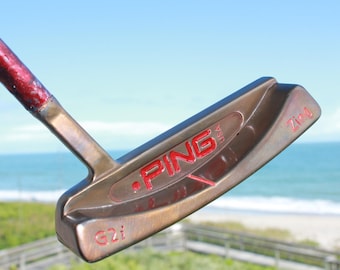 May include: A PING G2i Zing USA golf putter with a red and bronze finish. The putter has a red grip and the PING logo is prominently displayed in red. The background shows a beach and ocean.