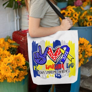 May include: White tote bag with a colorful graphic design. The design includes a peace sign, a heart, and a ribbon, with the text "Mom Daughter Dad Down Syndrome Awareness" and "3/21". The bag is carried by a person.