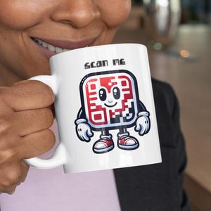 Op de afbeelding: Een witte keramische mok met een cartoon QR-code personage dat een mok vasthoudt met de tekst "Scan Me".