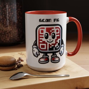 Op de afbeelding: Witte keramische mok met een rode rand en handvat. De mok heeft een cartoon QR-code met een glimlachend gezicht en de tekst "Scan Me".