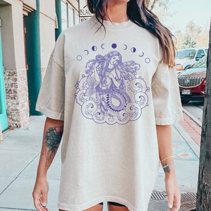 May include: A light beige oversized t-shirt with a purple graphic of a mermaid with long flowing hair, surrounded by clouds and a crescent moon design. The text "Life is tough, my darling, but so are you." is printed on the bottom of the shirt.