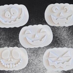 Puede incluir: Cinco cortadores de galletas blancos con forma de calabaza con diferentes caras talladas. Los cortadores est&aacute;n dispuestos sobre una superficie oscura espolvoreada con polvo blanco, creando un tema de Halloween.