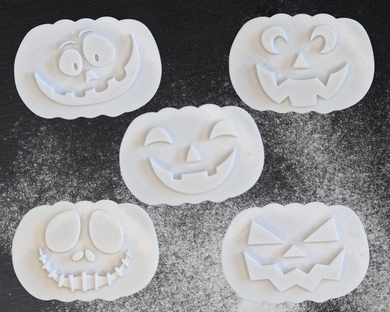 Puede incluir: Cinco cortadores de galletas blancos con forma de calabaza con diferentes caras talladas. Los cortadores est&aacute;n dispuestos sobre una superficie oscura espolvoreada con polvo blanco, creando un tema de Halloween.