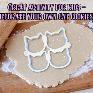 May include: A wooden rolling pin and a cat-shaped cookie cutter sit on a wooden cutting board with rolled-out dough. The text "GREAT ACTIVITY FOR KIDS - DECORATE YOUR OWN CAT COOKIES!" is displayed above the dough.