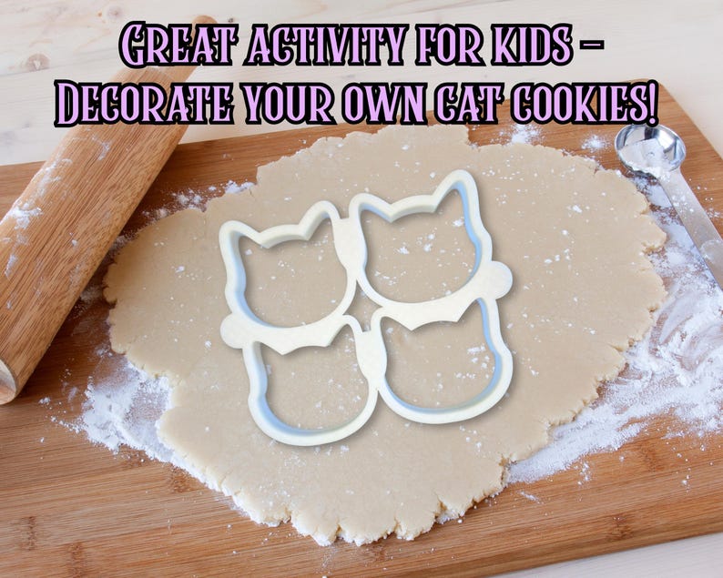 May include: A wooden rolling pin and a cat-shaped cookie cutter sit on a wooden cutting board with rolled-out dough. The text "GREAT ACTIVITY FOR KIDS - DECORATE YOUR OWN CAT COOKIES!" is displayed above the dough.