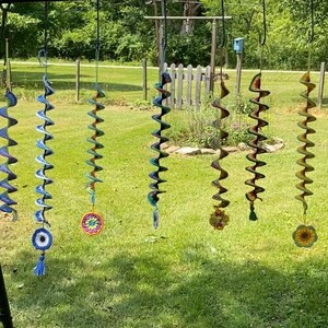 May include: A collection of handmade spiral wind chimes hanging outdoors. Each chime features a unique crochet design in various colors, including blue, green, and yellow. Some have decorative tassels or circular accents.