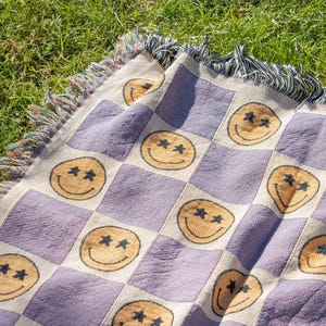 May include: A woven throw blanket with a checkerboard pattern of lavender squares and off-white squares. Each off-white square features a smiling face with star-shaped eyes in a golden-yellow color. The blanket has a fringed edge.