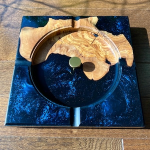 Puede incluir: Un cenicero redondo de madera y resina negra con un pomo de metal dorado. El cenicero tiene un diseño marmoleado azul y negro.