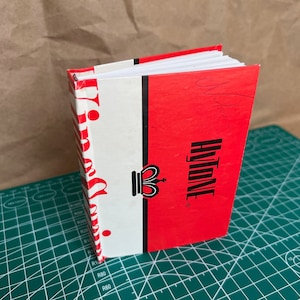 Può includere: Un quaderno rosso e bianco con una corona e la parola "HAYDENE" stampata sulla copertina.