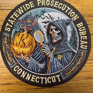 May include: A black and white embroidered patch with a grim reaper holding a magnifying glass and a jack-o-lantern. The text "STATEWIDE PROSECUTION BUREAU CONNECTICUT" is around the edge of the patch.
