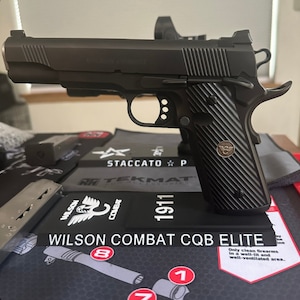 Könnte beinhalten: Eine schwarze Wilson Combat CQB Elite 1911 Pistole mit schwarzem Griff und silbernem Logo. Die Waffe liegt auf einer schwarzen und grauen Schießmatte mit dem aufgedruckten Text "STACCATO P" und "RATEKMAT".