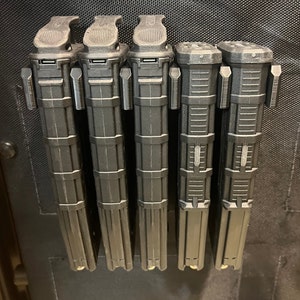 May include: Five black magazine holders with a textured surface and a clip on the top. The holders are attached to a black surface.