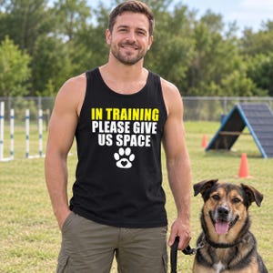 May include: A black tank top featuring the yellow and white text IN TRAINING PLEASE GIVE US SPACE above a white paw print graphic with a small heart in the center. The garment is worn with olive green cargo shorts and a black dog leash.