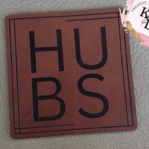 May include: A square, brown coaster with the word "HUBS" in large, black capital letters. The coaster has a thin, black border and is set against a gray surface. The upper right corner features a logo with the words "CREATIVE DESIGNS" and the letters "KL".