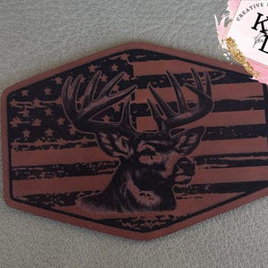 May include: A brown leather patch featuring a deer head silhouette with large antlers, set against a distressed American flag design. The patch has a unique shape and the text "CREATIVE DESIGNS KL" in the upper right corner.