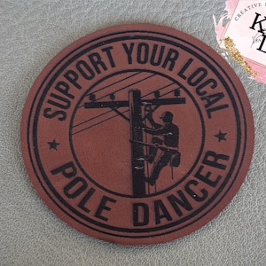 May include: A round, brown leather patch with the words "SUPPORT YOUR LOCAL POLE DANCER" in a circular design. The center features a silhouette of a lineman on a utility pole. The patch also has two stars.