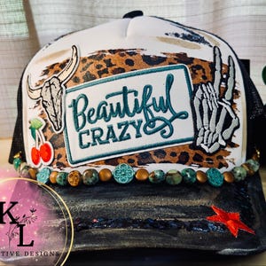 May include: A white and black trucker hat featuring a leopard print band, a central patch with the text "Beautiful CRAZY" in teal, a cow skull, a skeleton hand making a peace sign, a cherry graphic, and a row of turquoise and bronze beads.