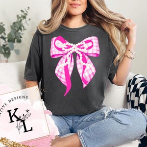 Puede incluir: Camiseta gris oscuro con un gran lazo rosa de cuadros vichy con detalles de perlas. El dise&ntilde;o del lazo est&aacute; centrado en la camiseta. La camiseta se combina con vaqueros azules. La imagen tambi&eacute;n incluye el texto "CREATIVE DESIGNS" y las letras "K L".