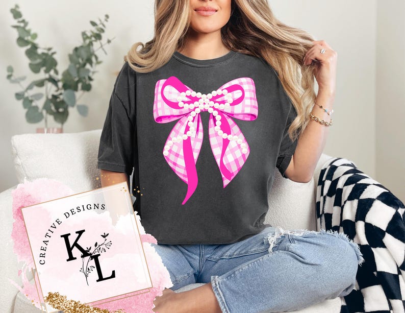 Puede incluir: Camiseta gris oscuro con un gran lazo rosa de cuadros vichy con detalles de perlas. El dise&ntilde;o del lazo est&aacute; centrado en la camiseta. La camiseta se combina con vaqueros azules. La imagen tambi&eacute;n incluye el texto "CREATIVE DESIGNS" y las letras "K L".